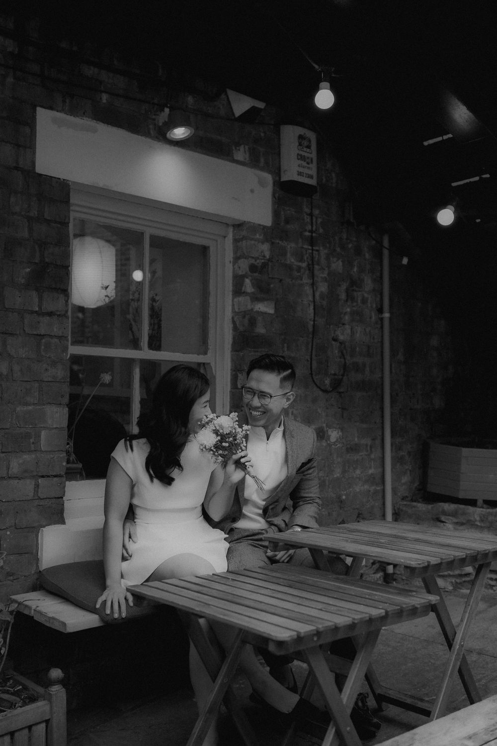 Black and white photo of a couple sitting at an outdoor cafe table, smiling at each other, with string lights and brick wall.