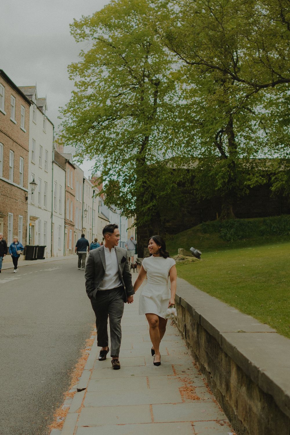 Couple walks hand-in-hand along a cobblestone street in a historic town, woman in white dress, man in gray suit.