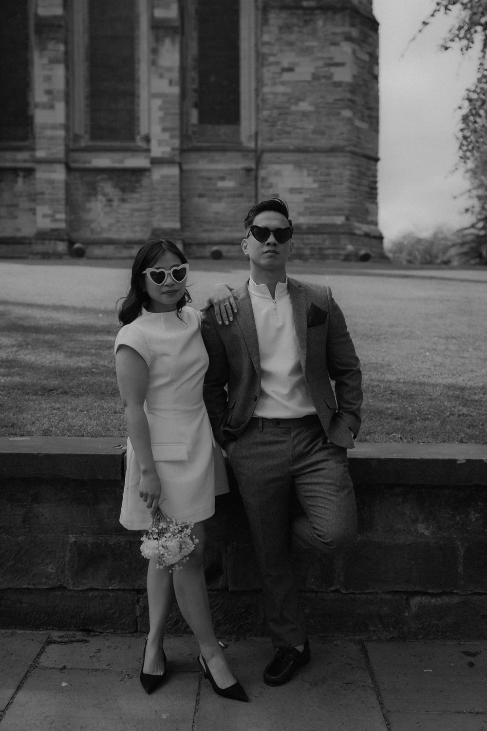 Stylish couple in black and white, posing in sunglasses outside a historic stone building on their wedding day.