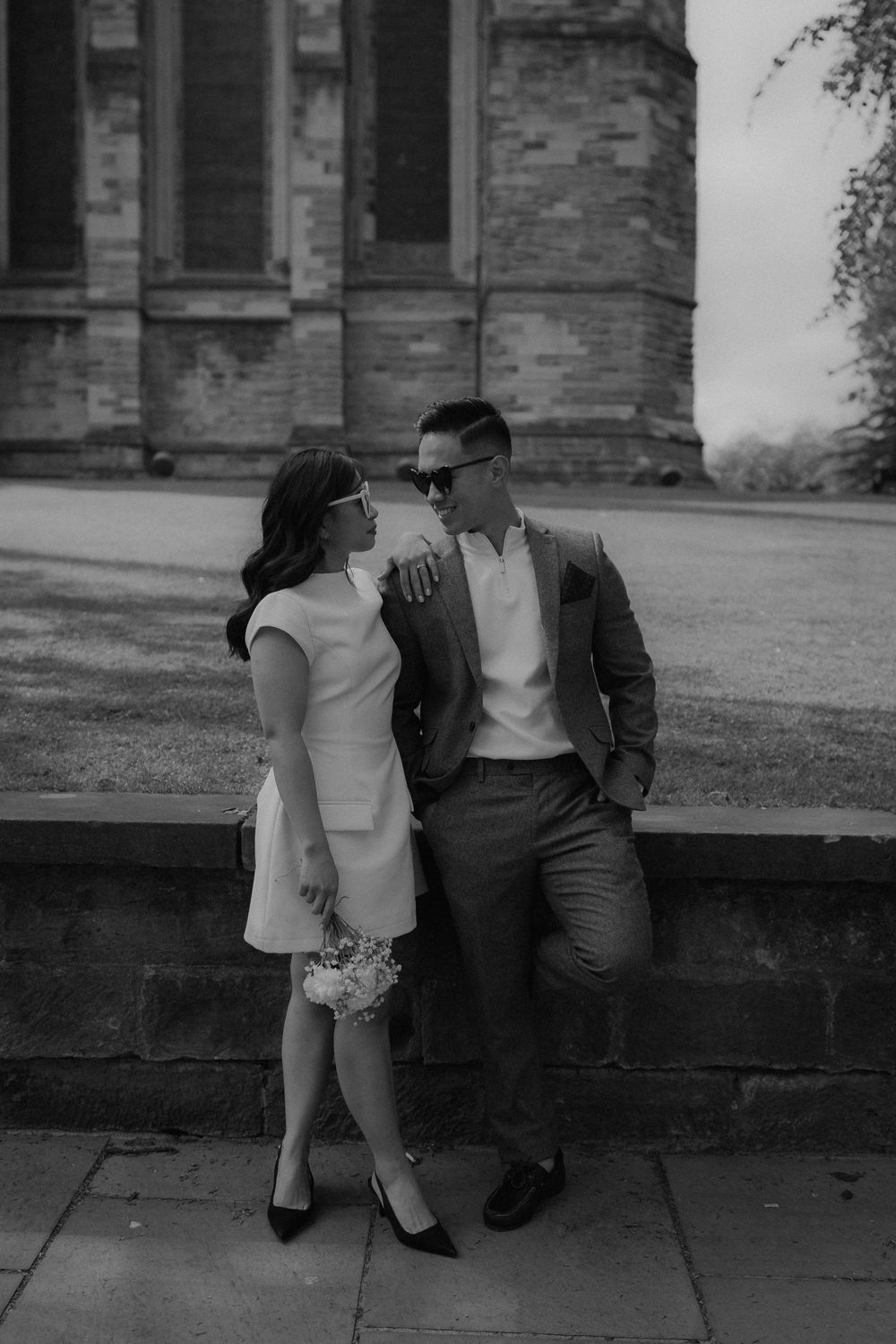 Black and white photo of an elegant couple posing near a stone wall outside a historic brick building.