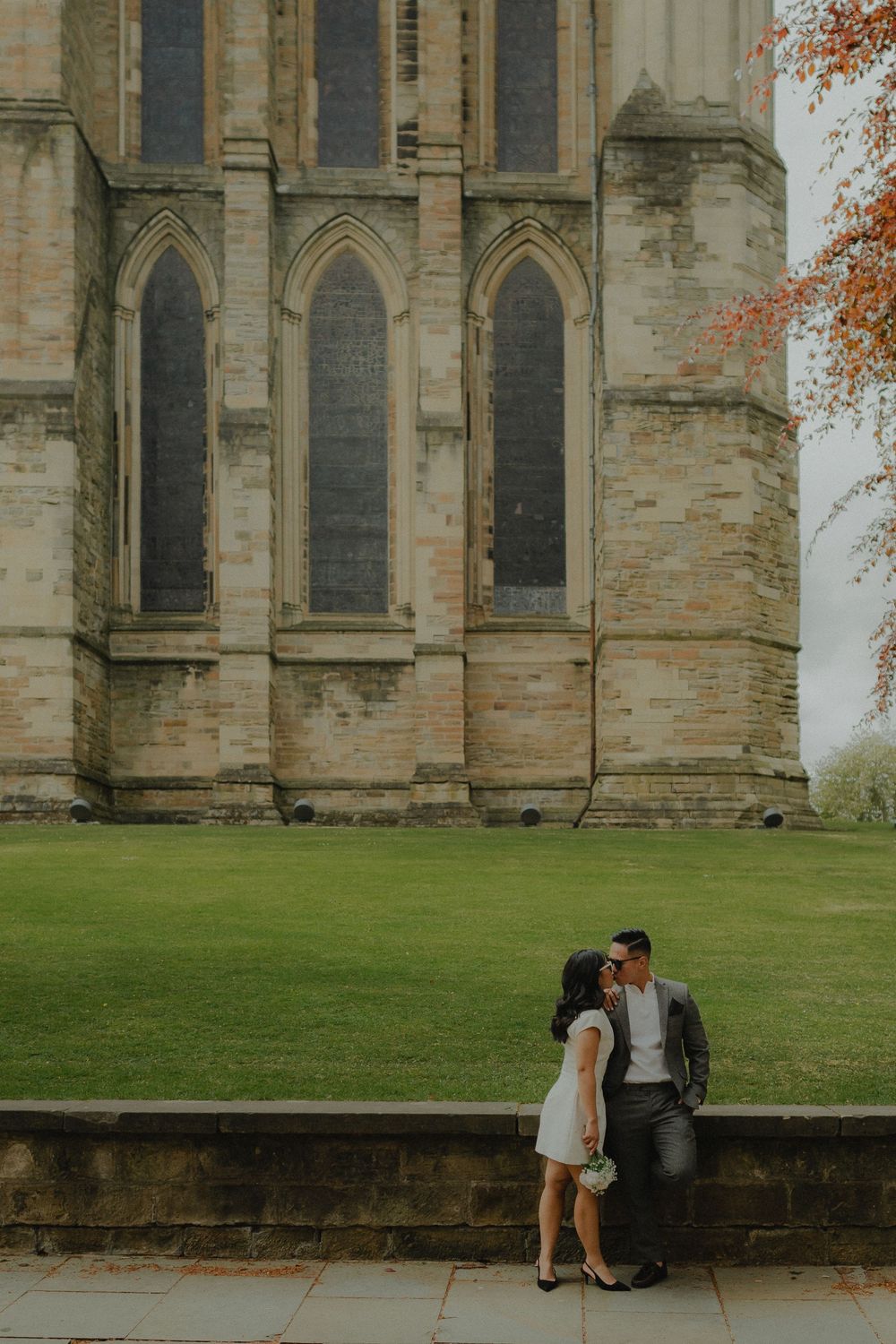 Couple sharing a romantic kiss outside a grand Gothic stone cathedral with tall arched windows and lush green lawn.