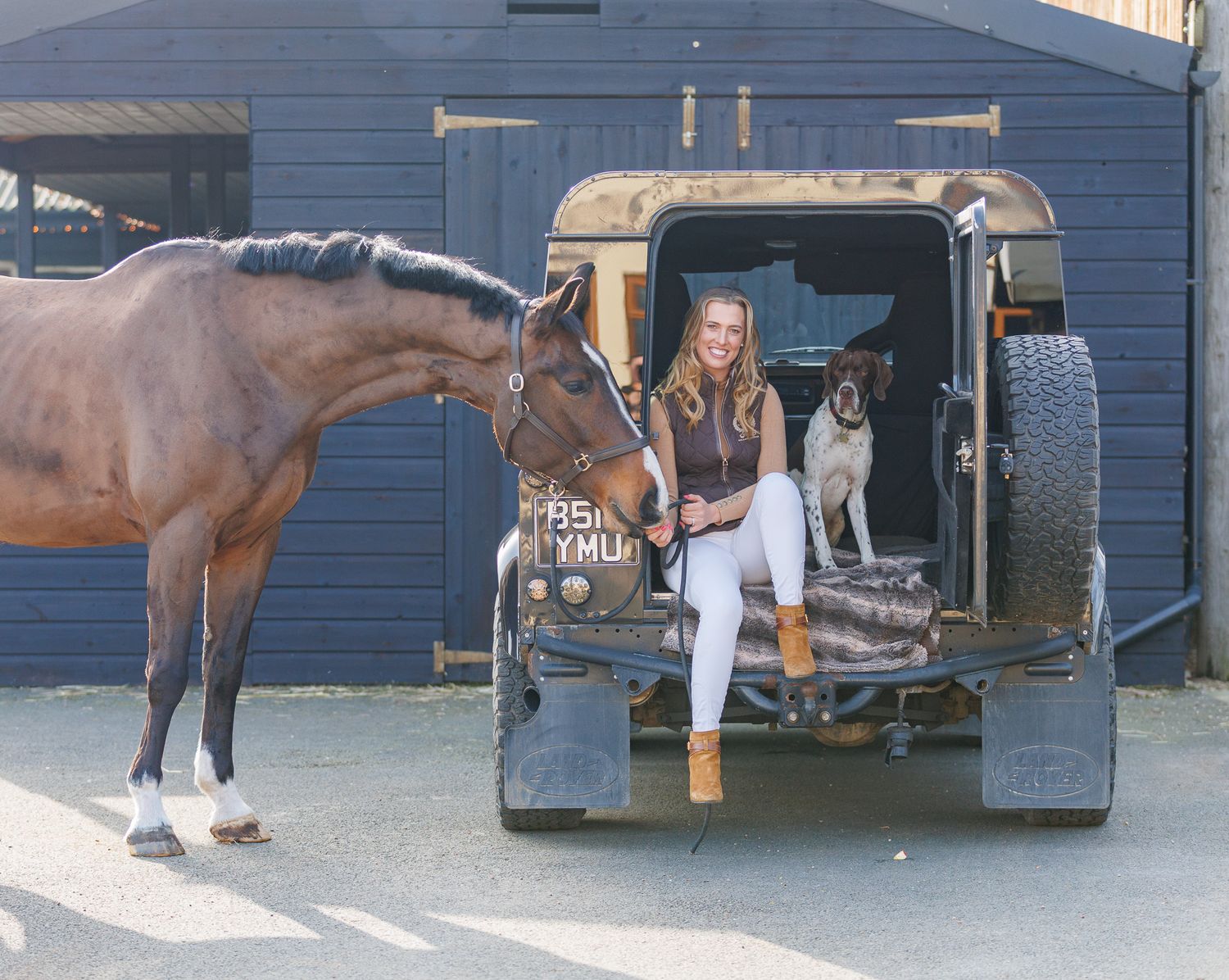 Two riders stand with their animals - a dog and a dark horse relaxing outdoors in a black and white photo.