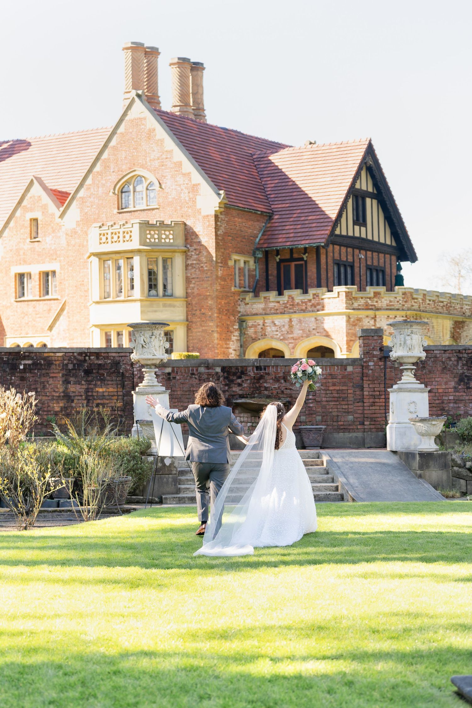 Bride and groom walking hand in hand across green lawn toward elegant brick Tudor-style mansion wedding venue.