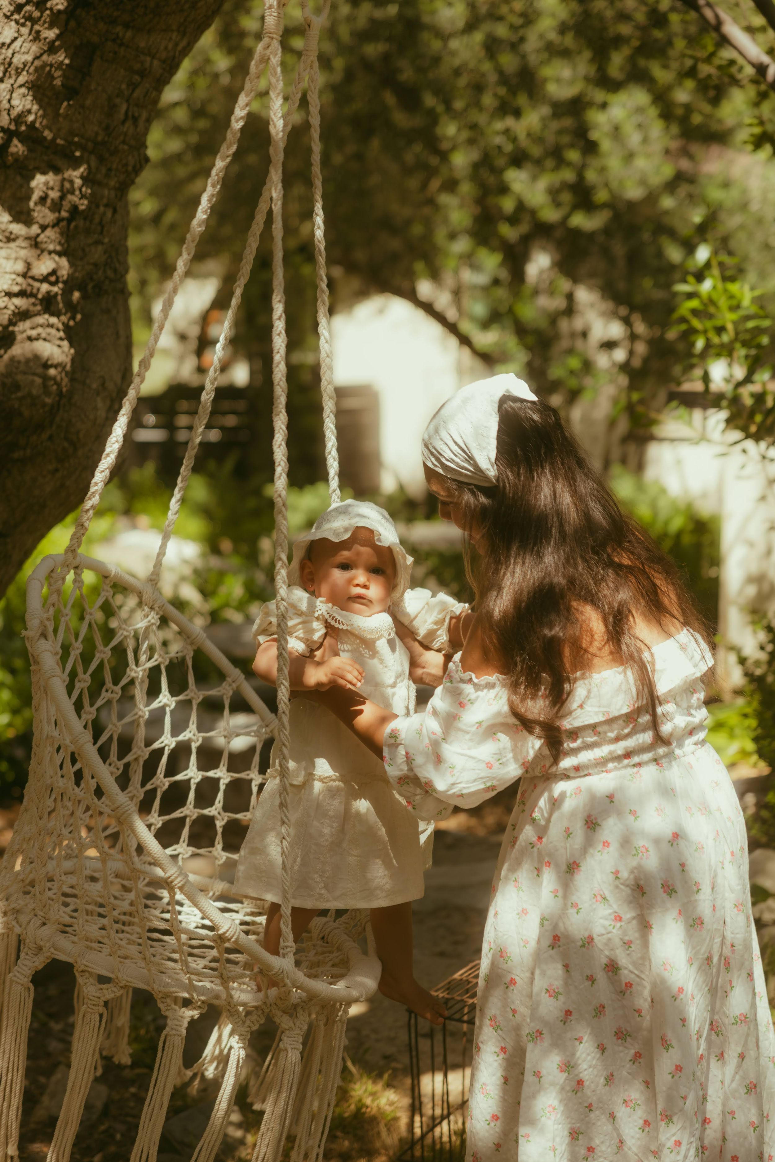 Mother in floral dress holds baby in white bonnet beside swing chair in sunny garden for a mini session with Yoyo Leung, LA photographer