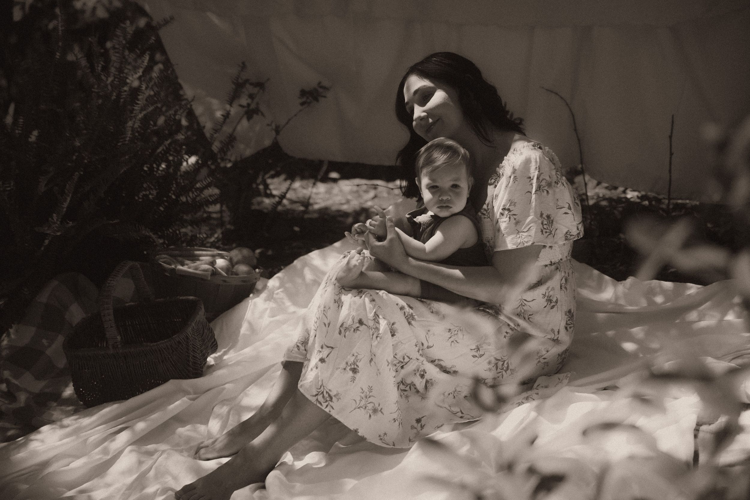 Black and white photo of a mother cradling her baby outdoors, sitting on a flowing white dress in a garden.