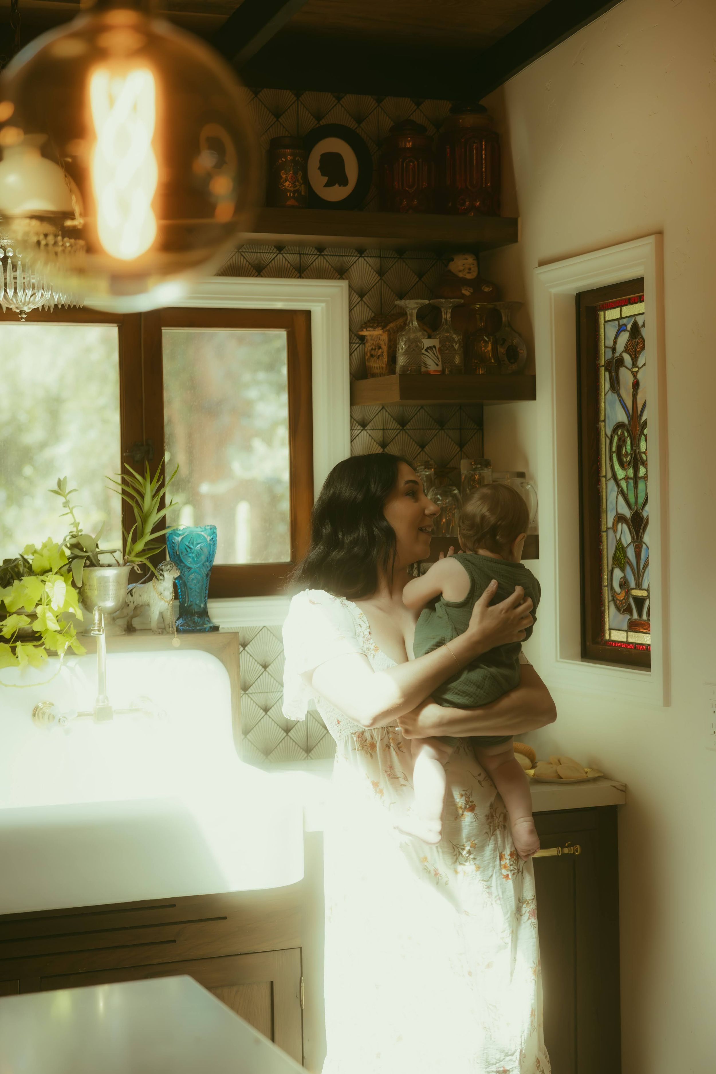 A woman in a white dress tenderly kisses a baby she holds in a warmly lit, vintage-style kitchen.