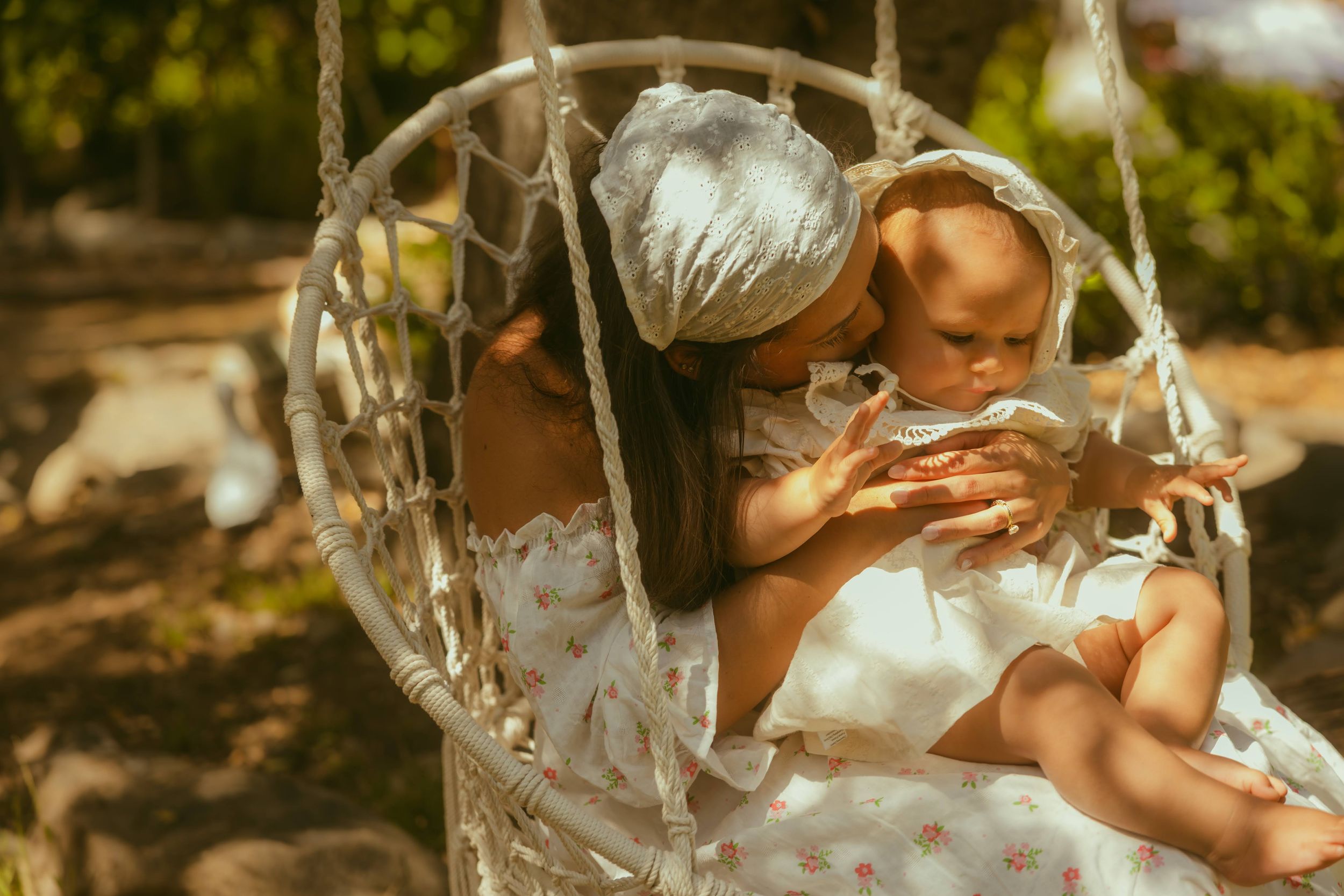 Mother in white floral dress kissing baby in hanging macrame chair outdoors in sunny garden.