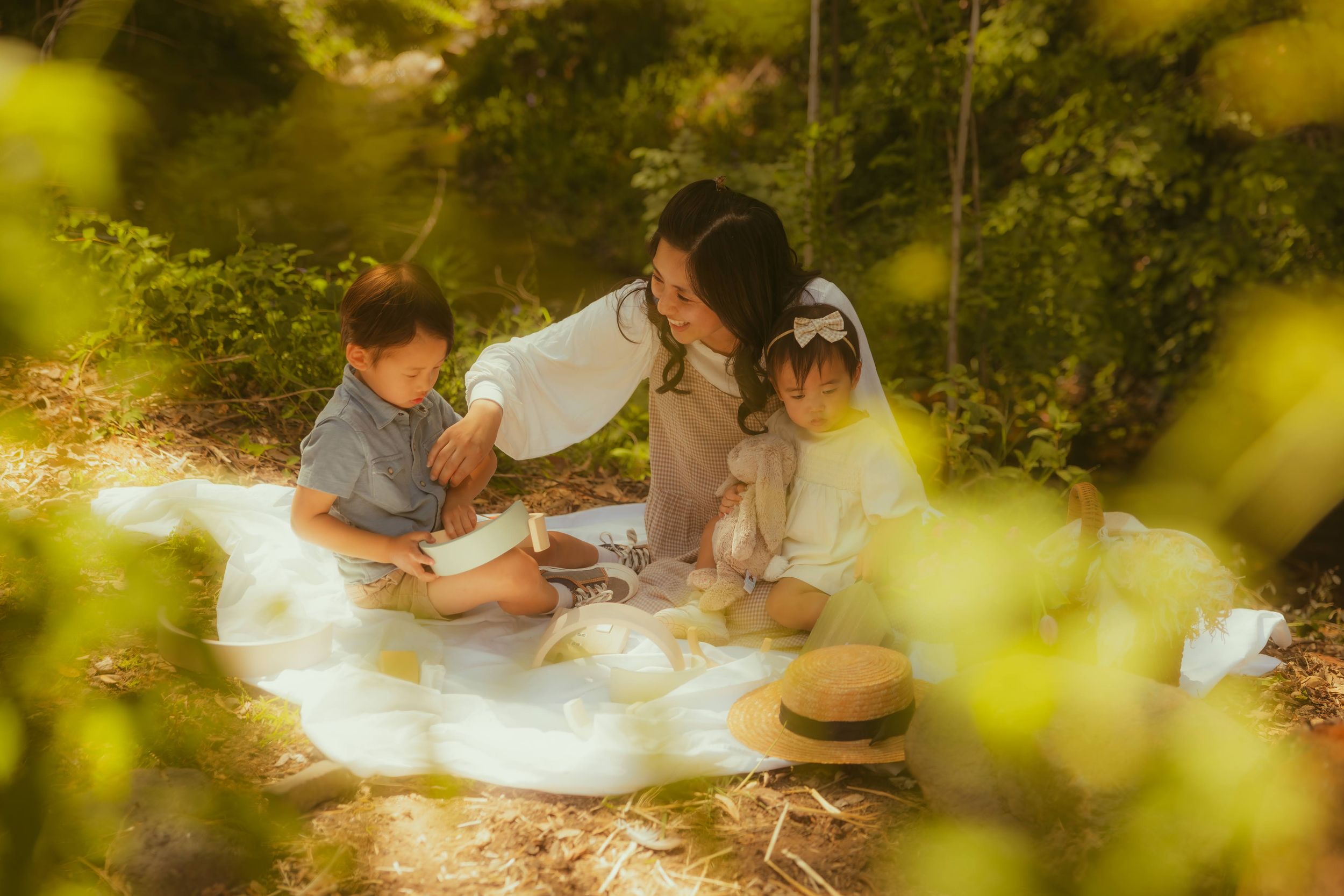 A mother and two young children enjoy a peaceful outdoor picnic on a white blanket surrounded by lush green trees.