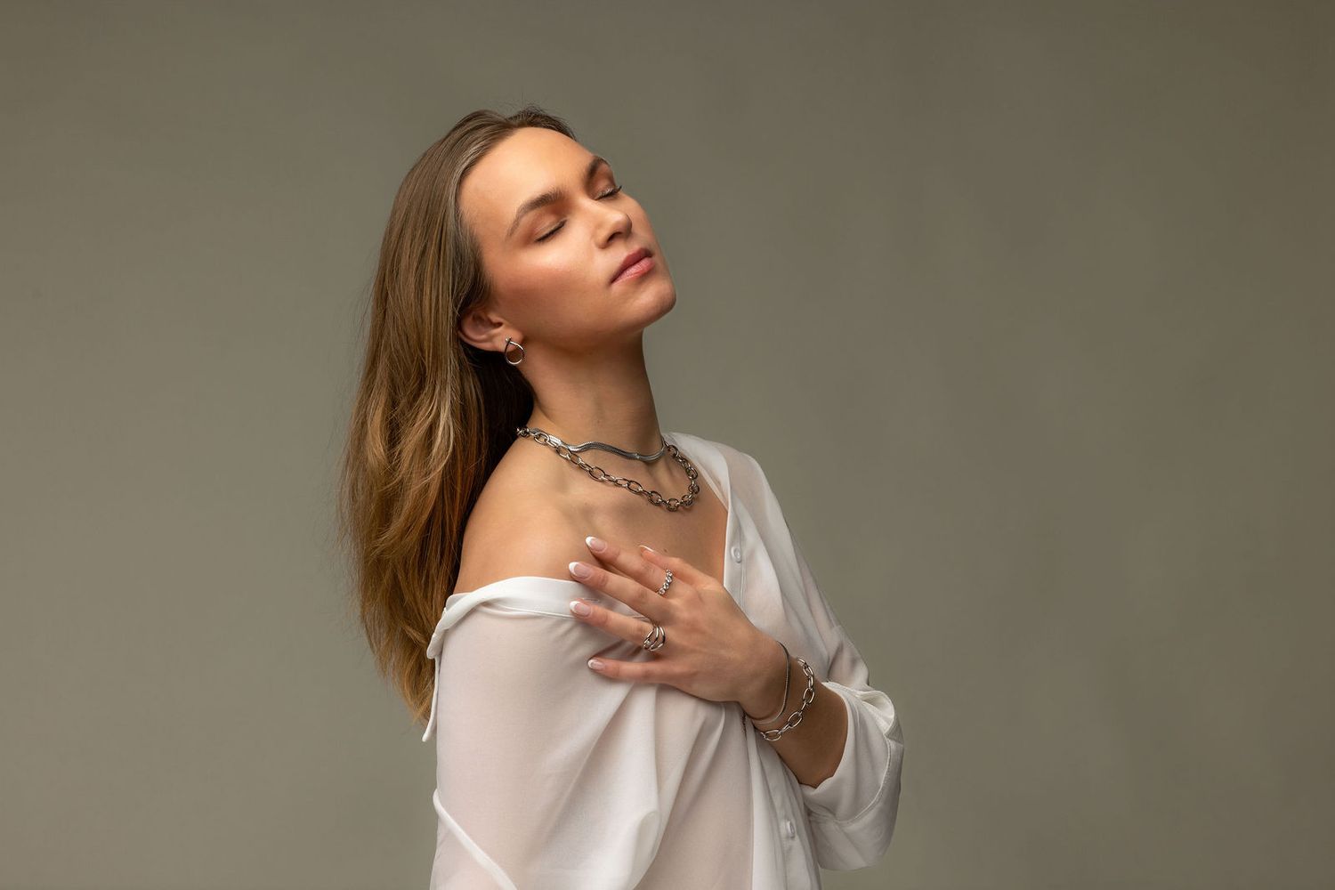 Woman in white off-shoulder blouse wearing silver jewelry, posing gracefully against a gray background.