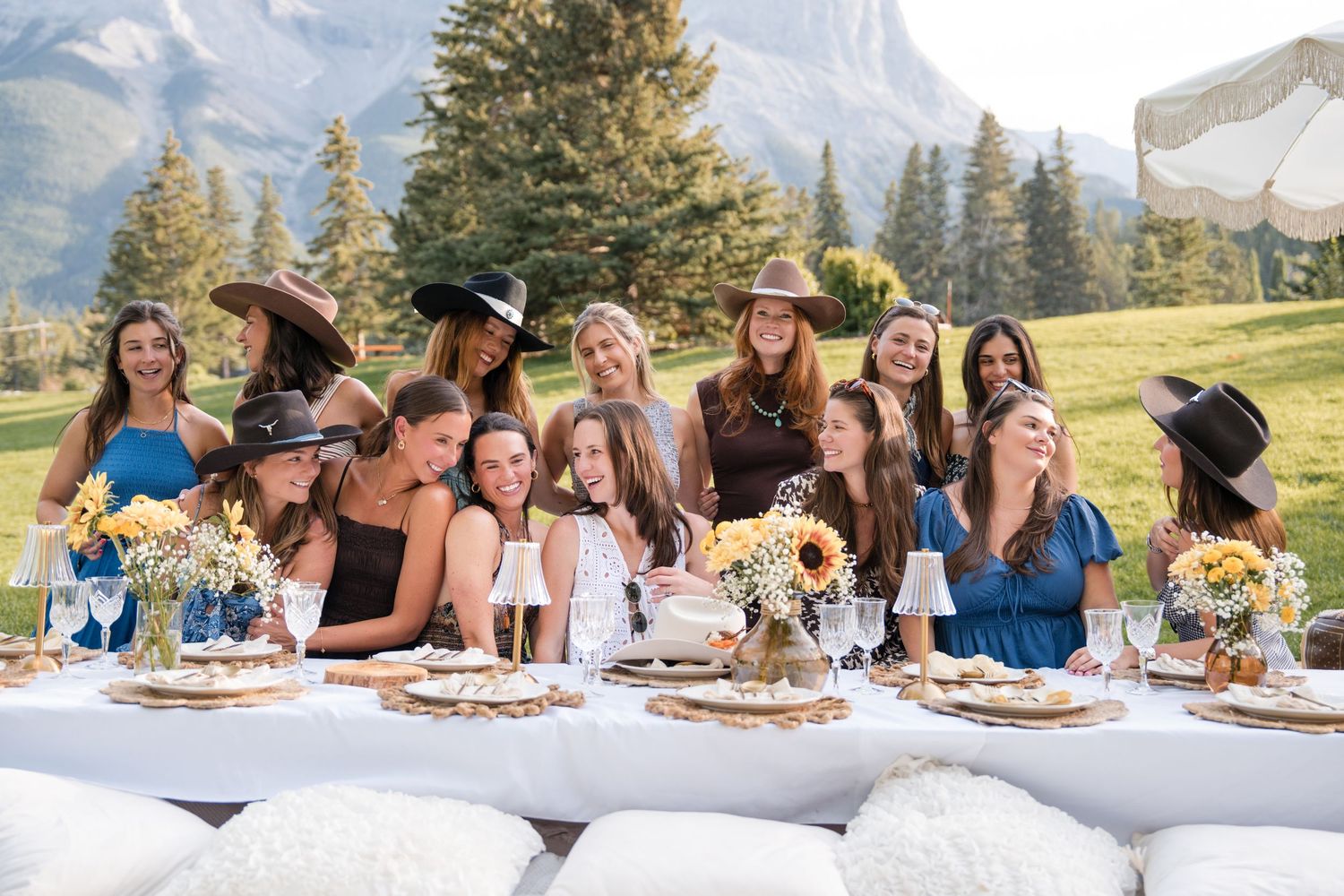 Group of women in hats laughing at an outdoor mountain birthday party table with floral centerpieces in Canmore, Alberta.