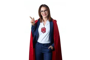 Woman in red superhero cape and glasses smiling, wearing a business suit with a logo badge on white background.
