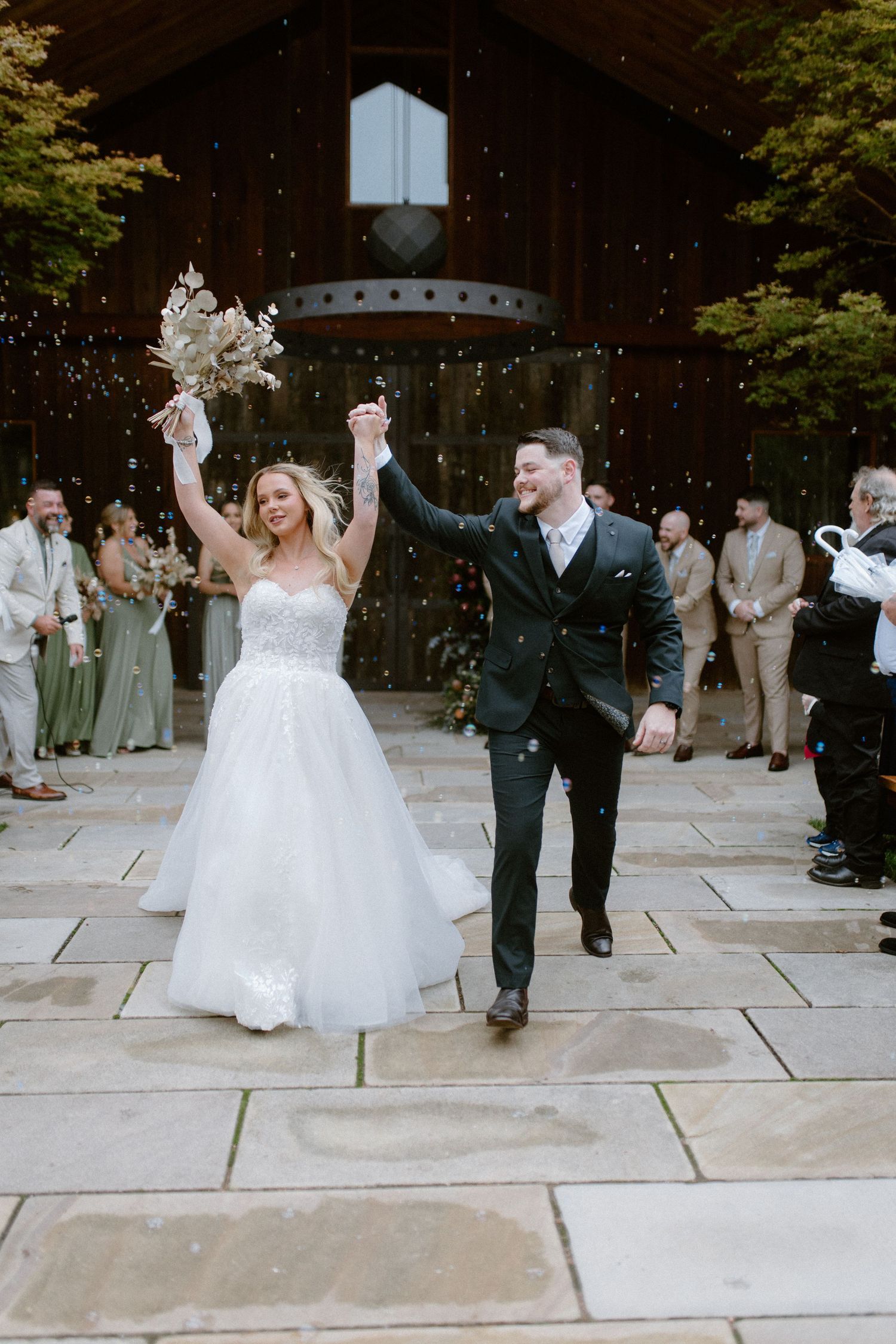 Joyful newlyweds celebrate with confetti outside a rustic barn venue, bride holding bouquet aloft.