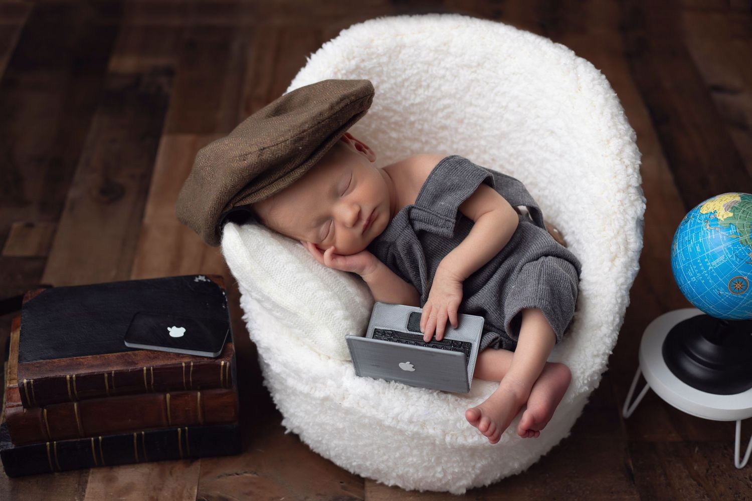 Newborn baby boy lies comfortably in a white papasan chair while using a laptop computer with Iphone with paperboy hat with books and globe.