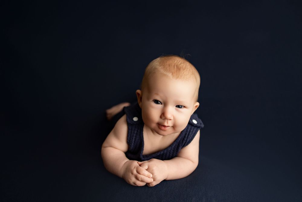 Adorable baby in navy romper lying on tummy against dark background, smiling at camera during professional photo session.