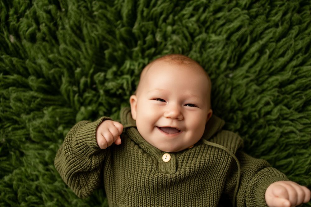 Smiling baby in green knit sweater lying on green fluffy rug, looking up with a joyful expression.