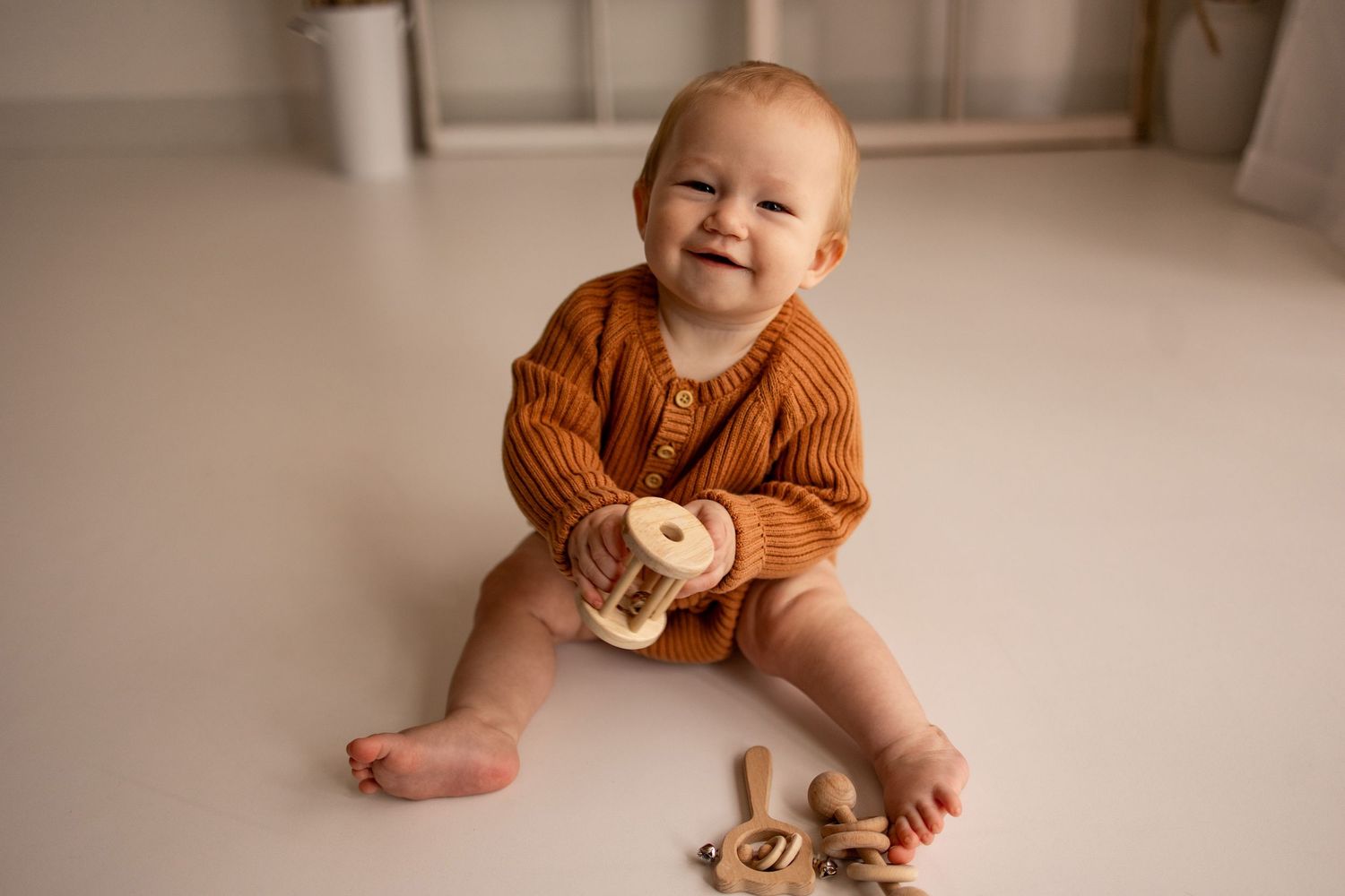 Smiling baby in rust knit romper sits on white floor playing with wooden stacking toy.