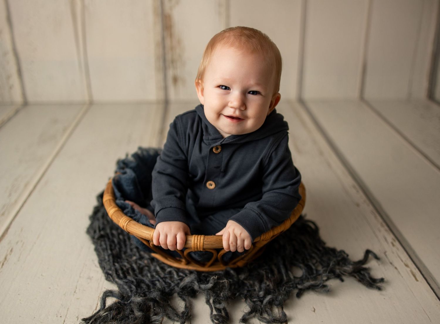 Smiling red-haired baby in navy outfit sitting in a wicker bowl on a white wood floor with blue knit blanket.