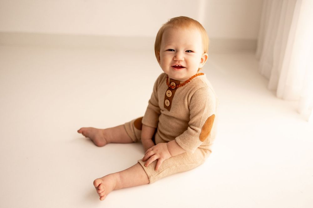 Smiling baby in a beige knit romper sitting on a white floor in a bright, airy studio setting.