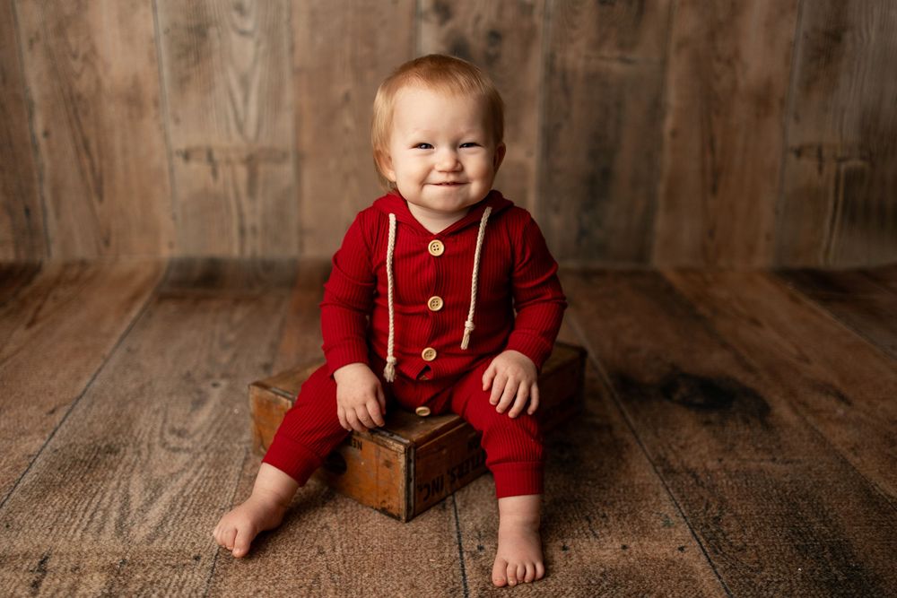 Smiling baby in red romper sitting on wooden box against rustic wood backdrop in professional photo.