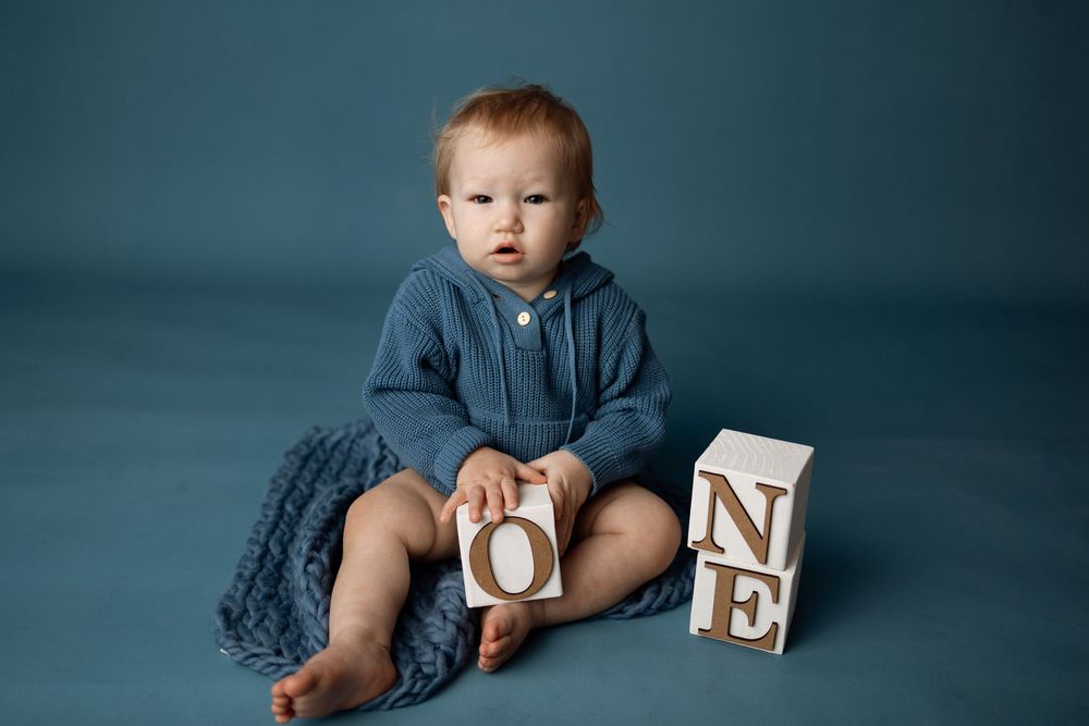 Baby boy in blue romper sits with wooden ONE blocks on blue backdrop, celebrating his first birthday milestone.