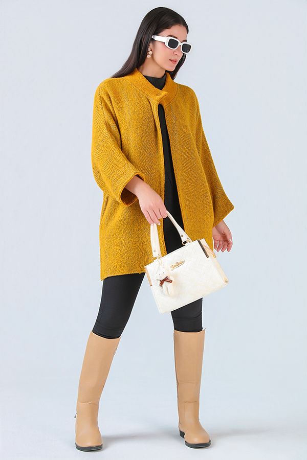Woman in mustard yellow knit cardigan, black outfit, beige boots, and white handbag poses against light blue background.