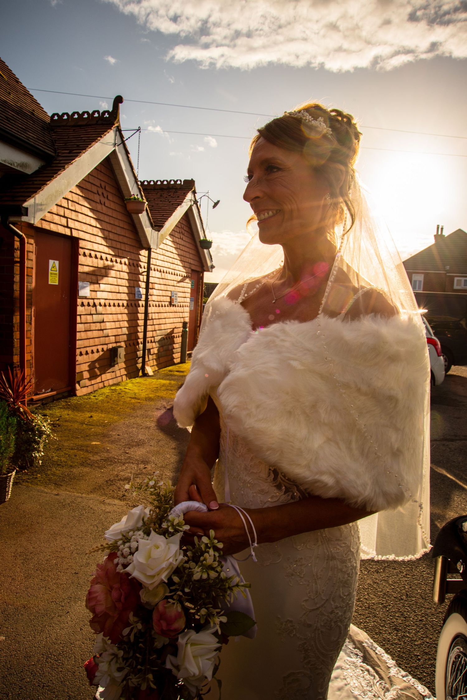 Radiant bride in white lace gown and fur shawl holds bouquet outdoors, backlit by golden sunset near rustic buildings.