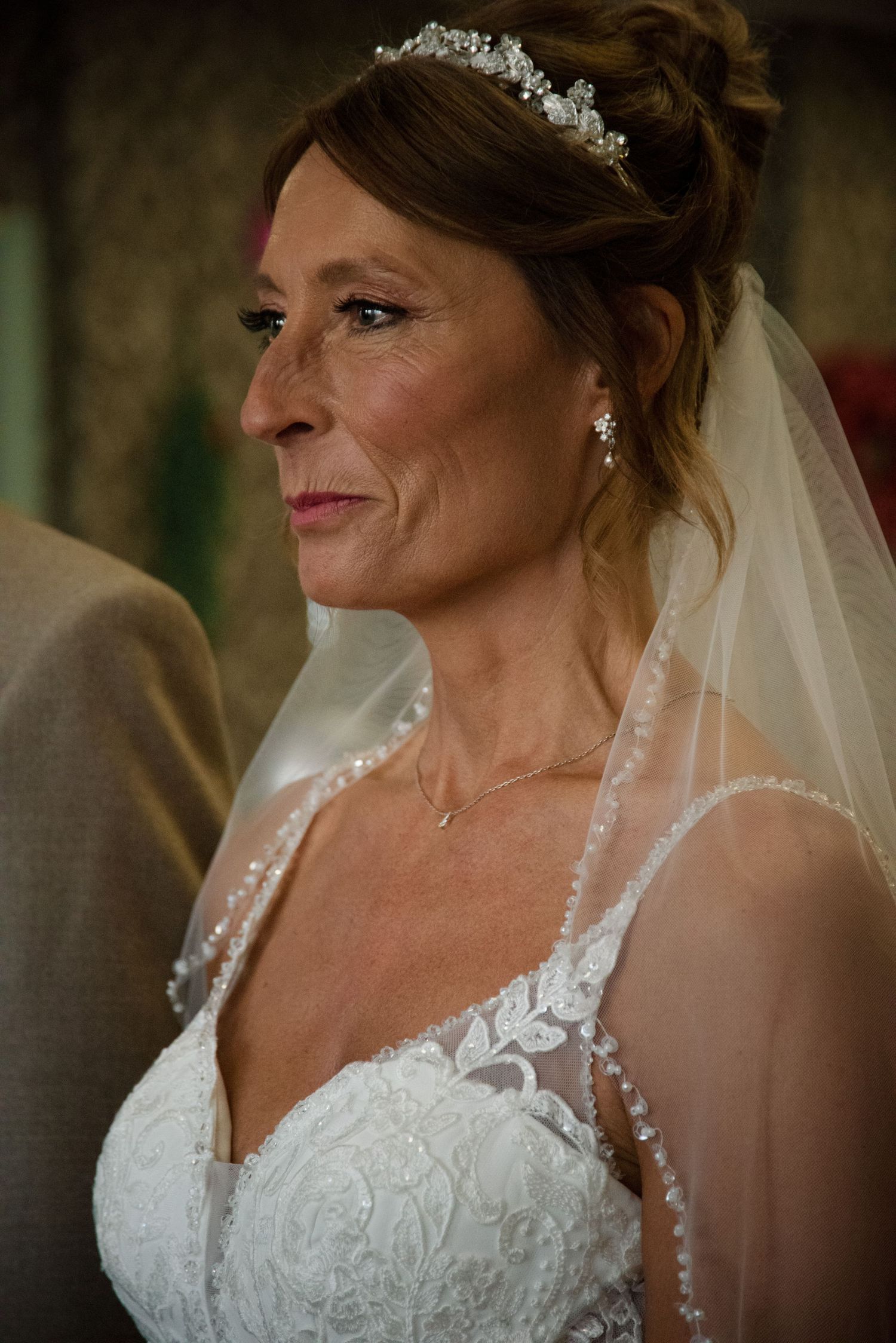 Elegant bride in lace wedding dress wearing pearl tiara and veil, smiling softly during wedding ceremony.