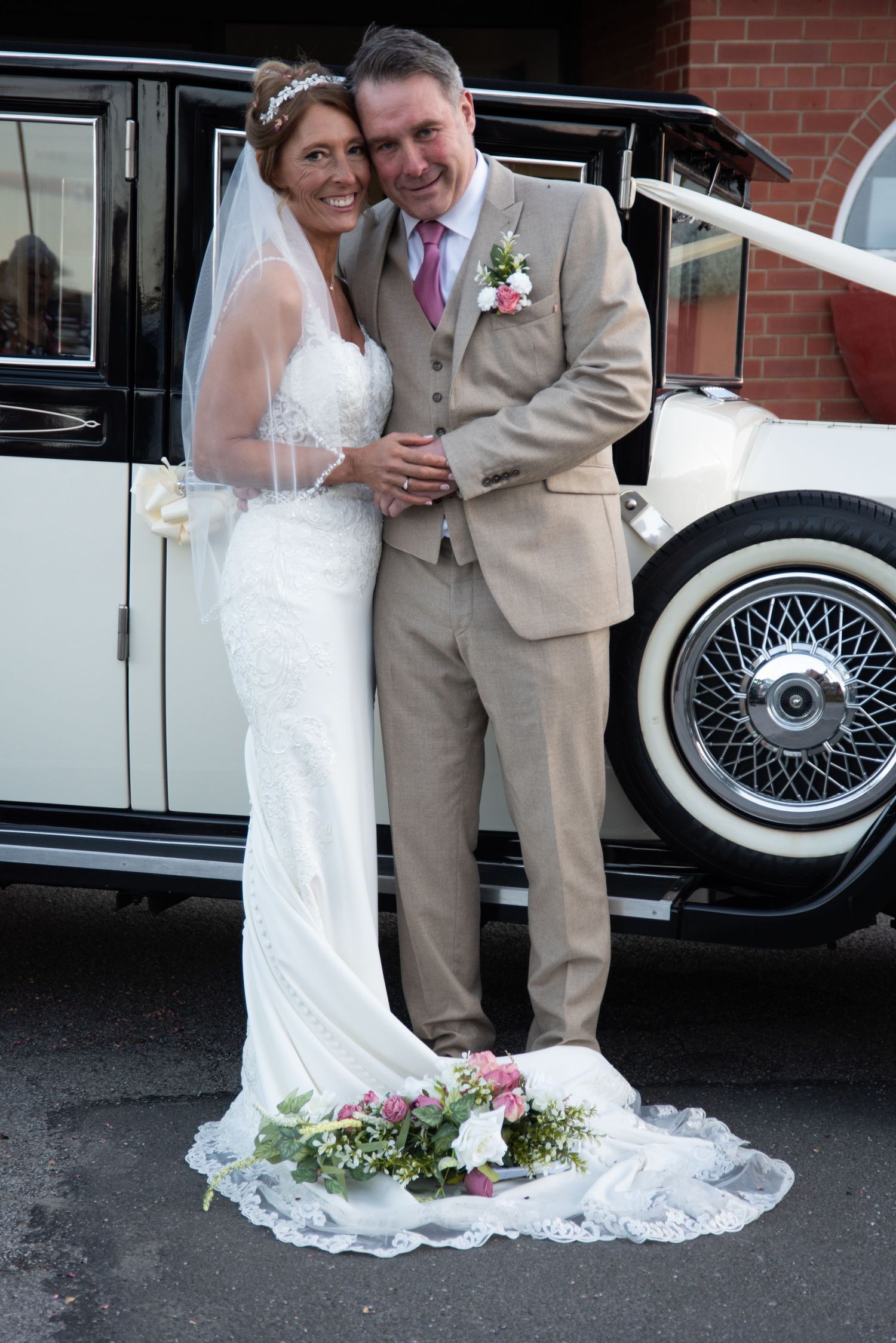 Bride and groom pose beside a classic vintage wedding car, bride in lace gown with veil and bouquet.