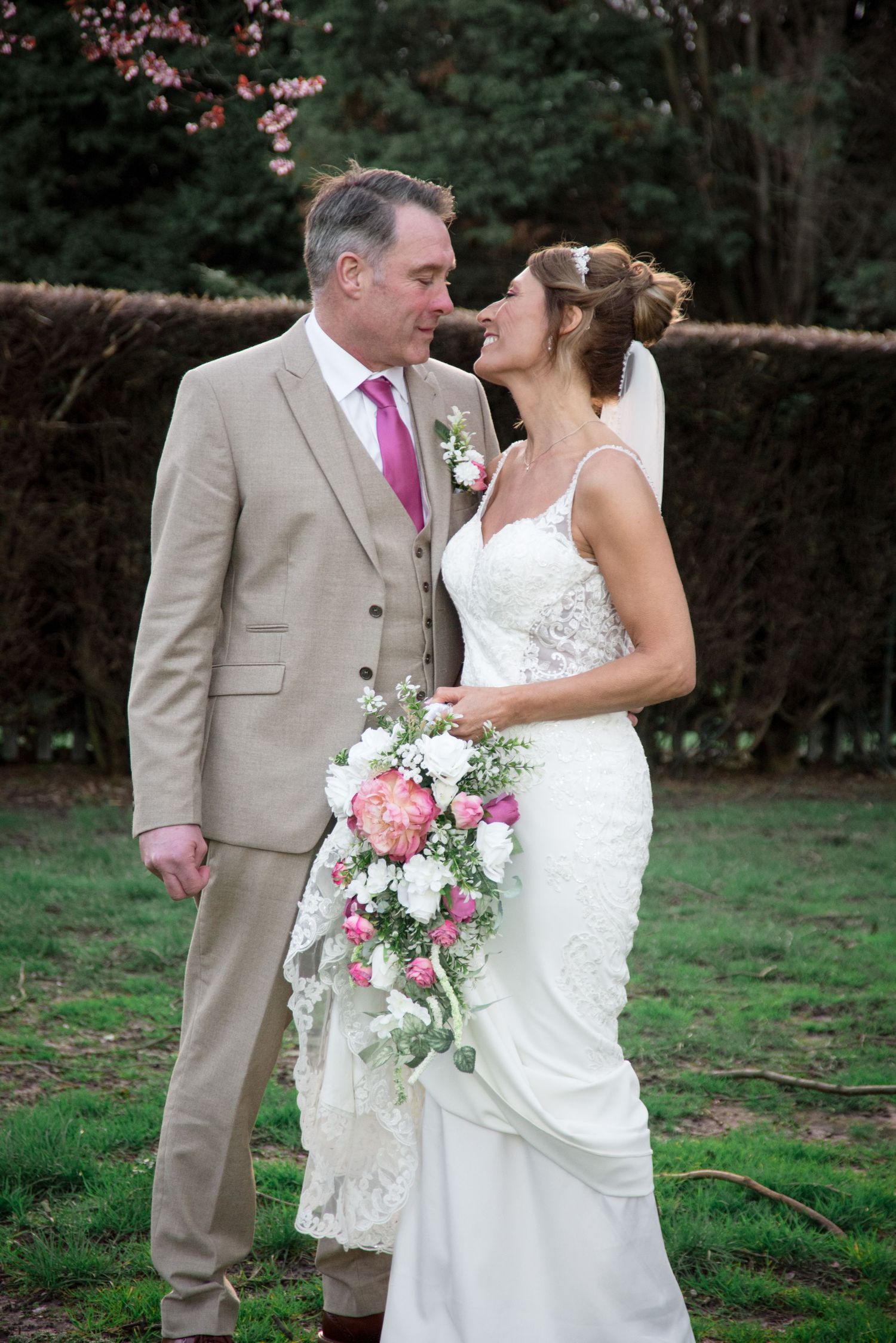 Bride and groom sharing a romantic moment outdoors, bride holding pink and white cascading bouquet, groom in tan suit with pink tie.
