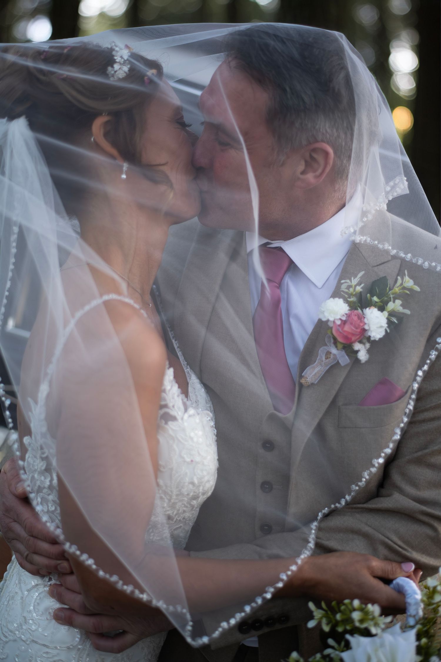 Bride and groom sharing a romantic kiss under a white wedding veil, groom wearing tan suit with pink tie.