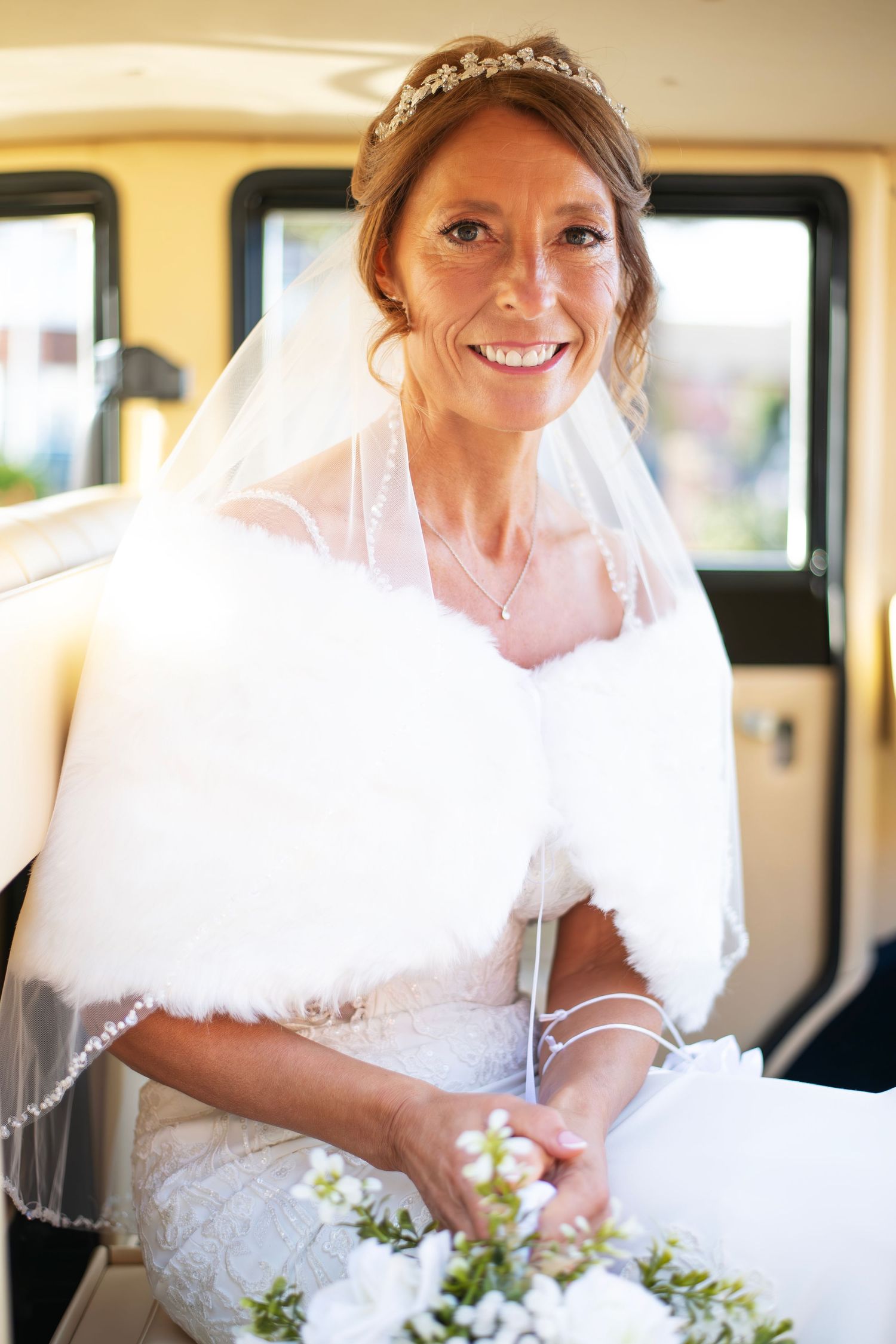 Smiling bride in white lace dress, fur wrap, and tiara with veil holds bouquet inside a vintage car.