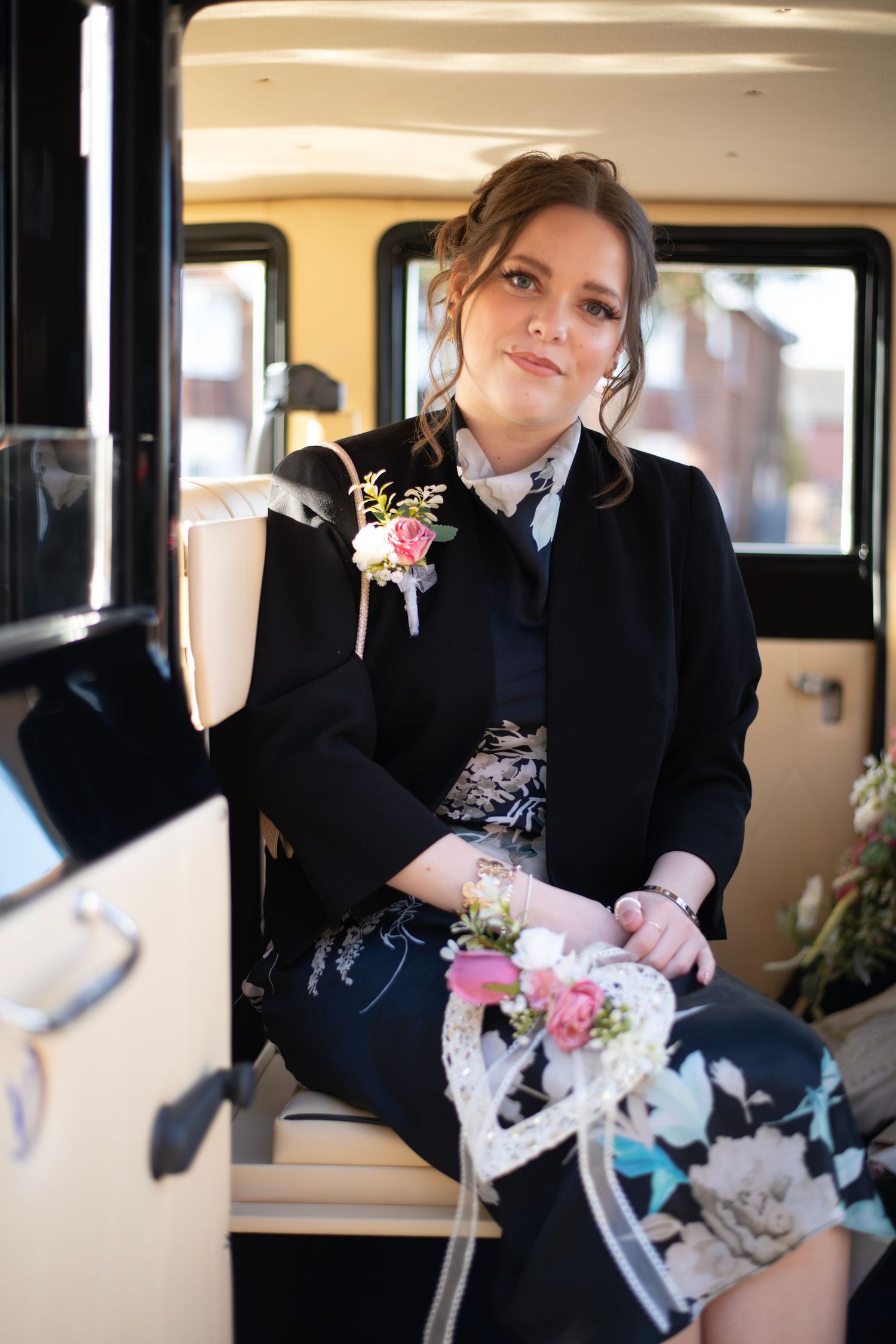 Young woman in black jacket with floral corsage sits inside a vintage wedding car holding a decorative heart.