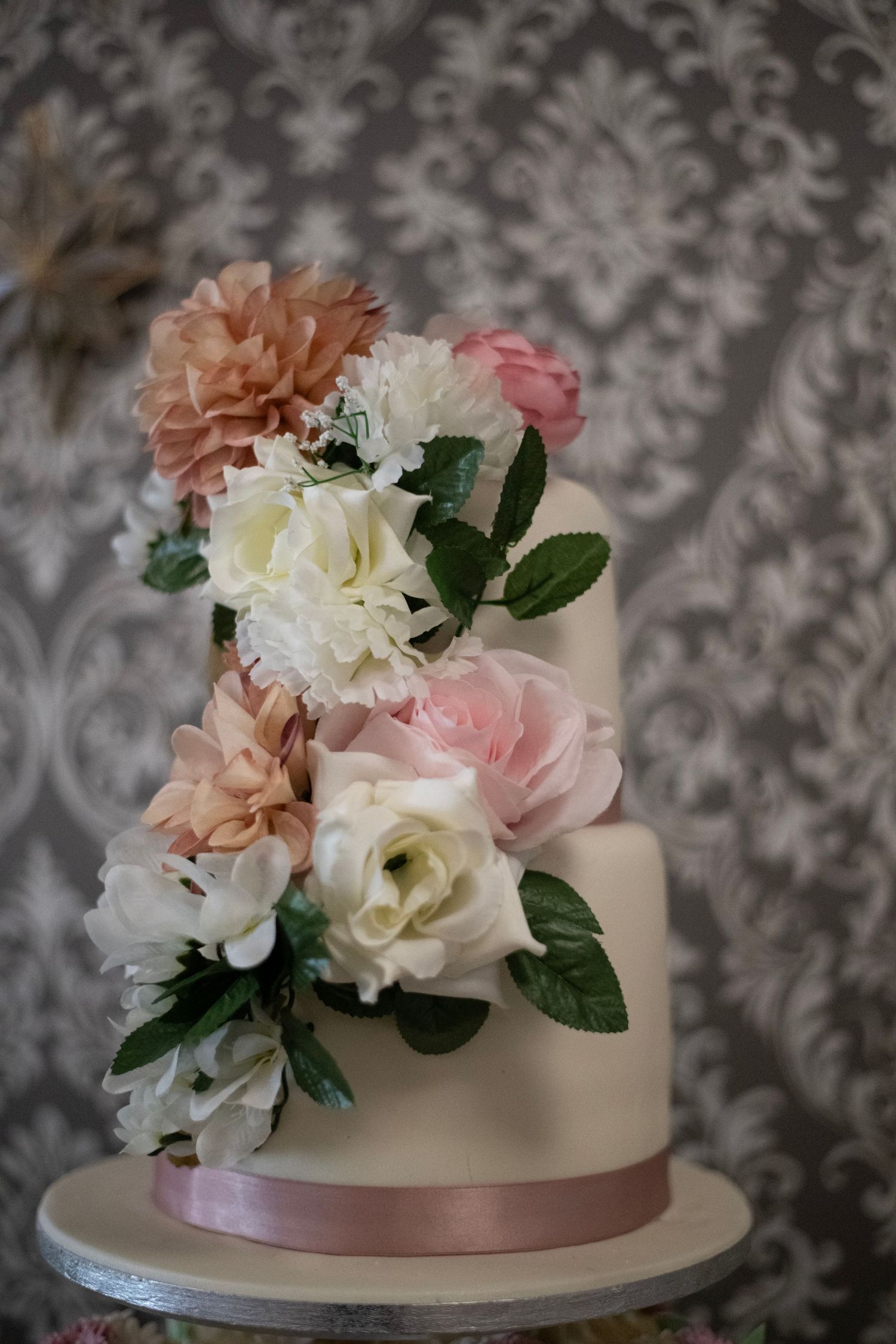 Elegant three-tier white wedding cake adorned with cascading pink, peach, and cream sugar flowers and green leaves.