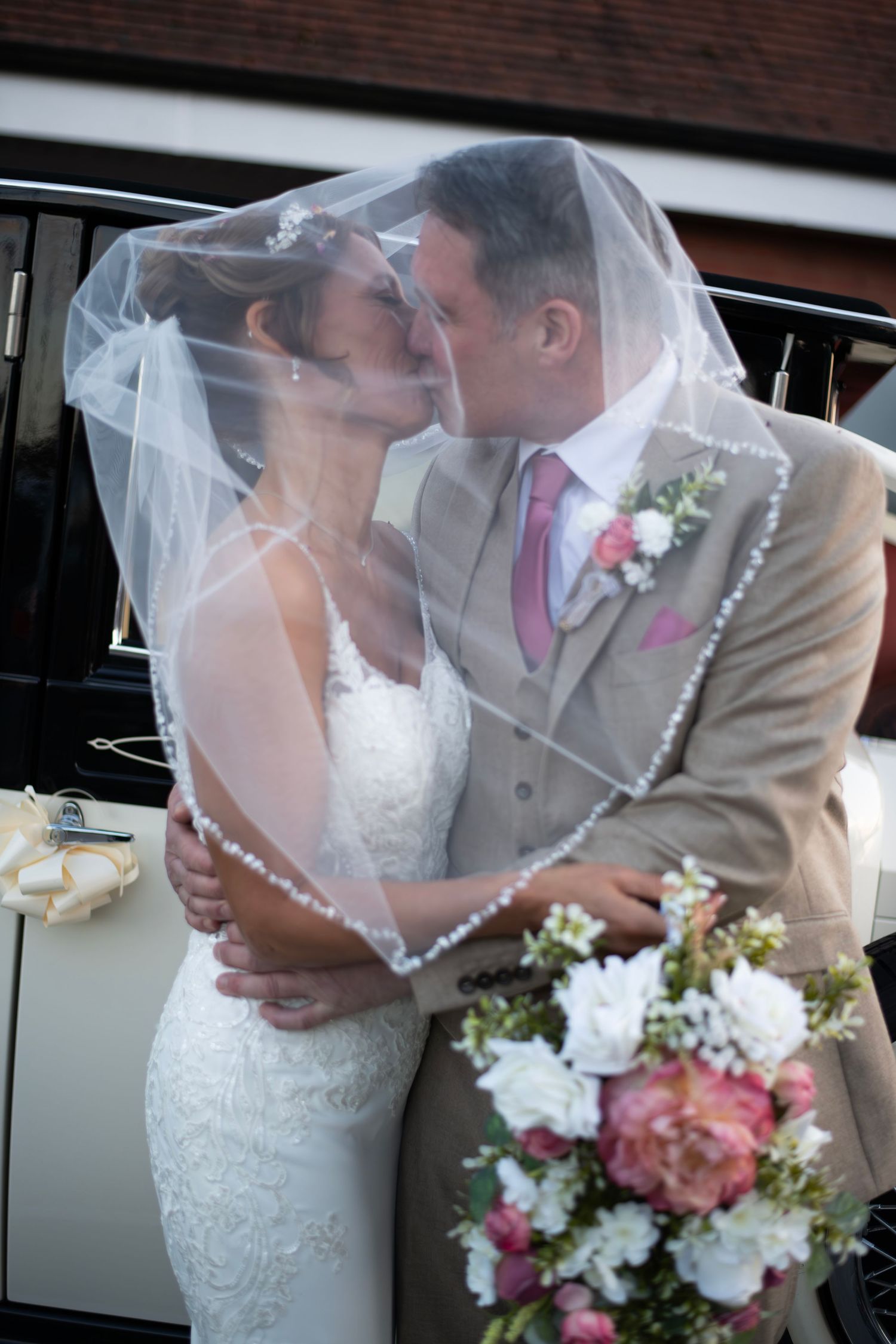 Bride and groom sharing a romantic kiss under a beaded wedding veil, bride holding pink and white bouquet by a vintage car.