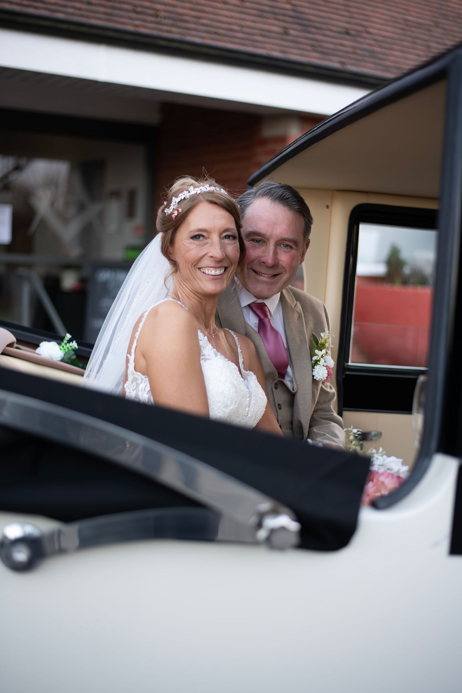 Smiling bride and groom pose together inside a vintage white wedding car, bride wearing veil and lace dress.