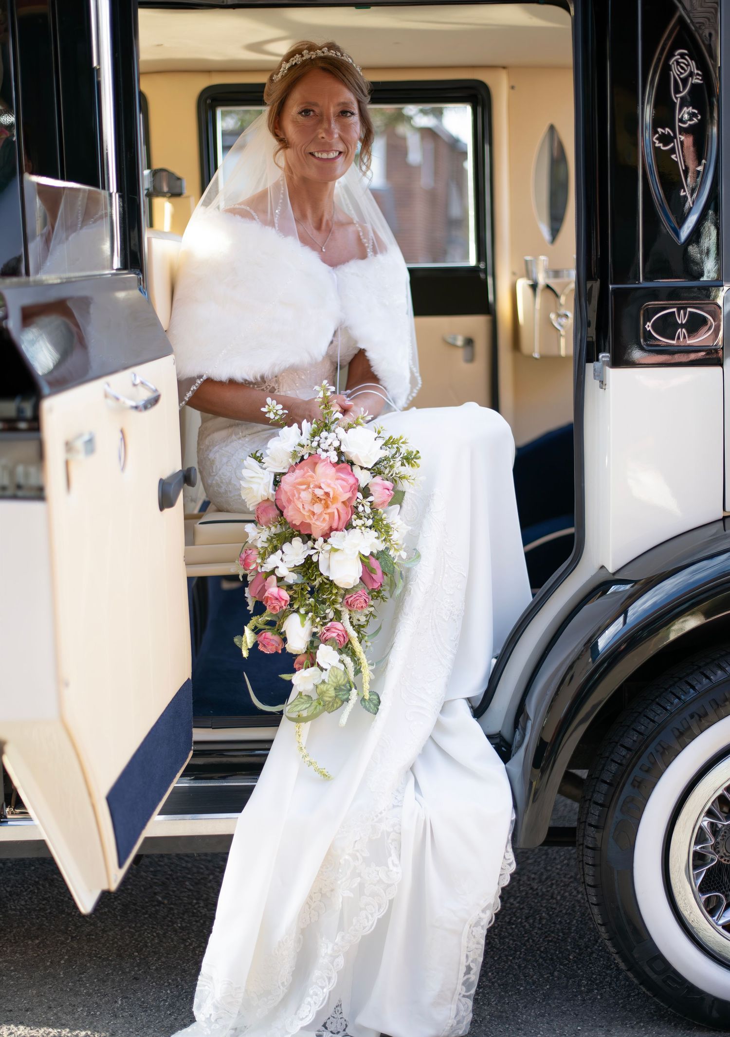 Smiling bride in white gown, fur stole, and veil holds cascading pink bouquet while stepping out of vintage wedding car.