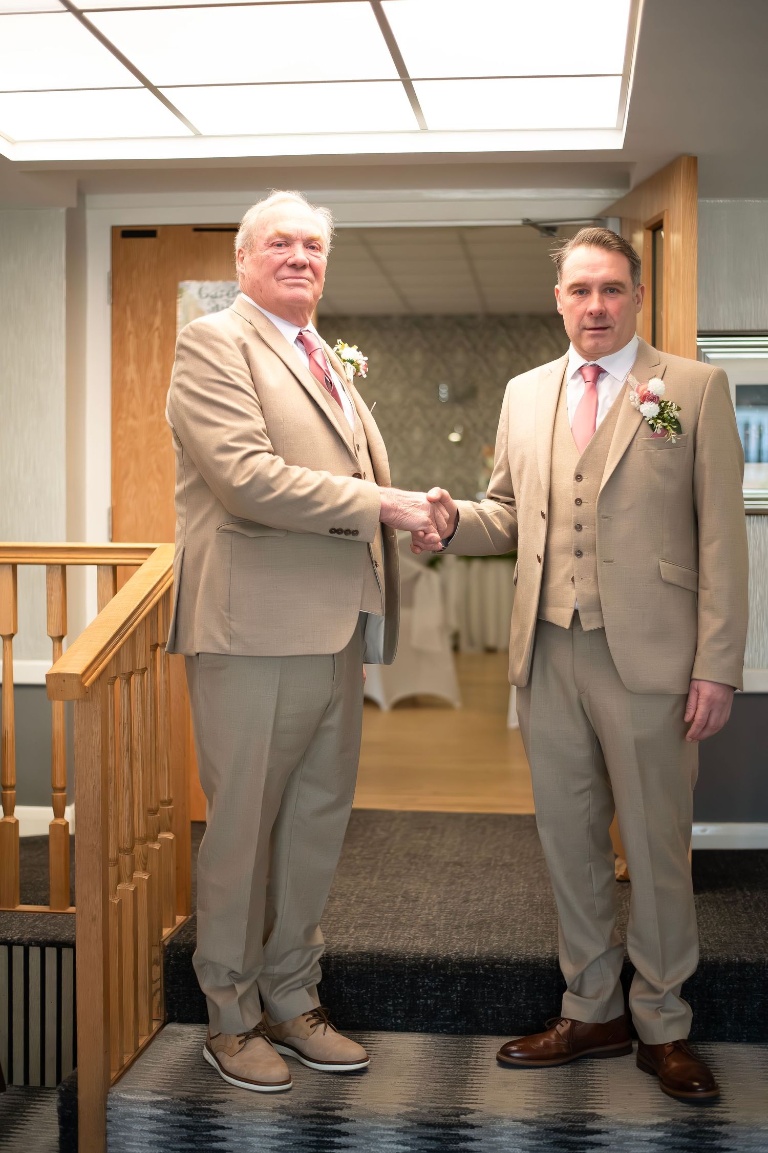 Two men in tan suits shaking hands at a wedding venue, both wearing boutonnieres and formal attire.