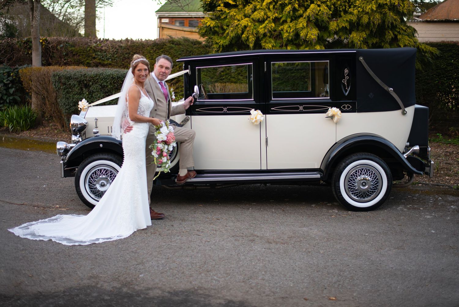Bride and groom posing beside a vintage black and cream wedding car decorated with white ribbons.
