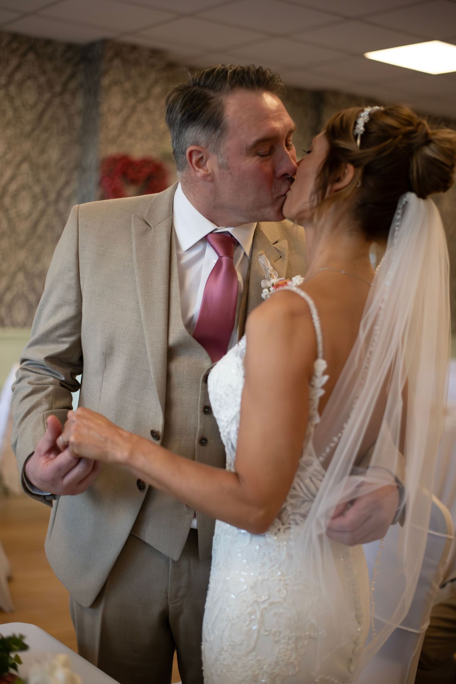 Bride and groom sharing a first kiss at their wedding reception, groom in tan suit with burgundy tie.
