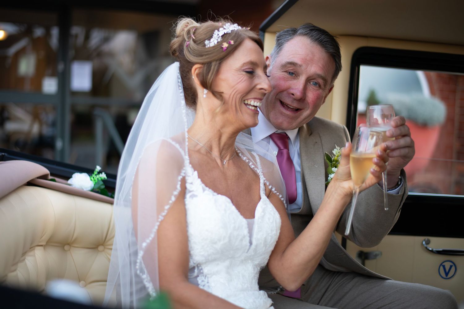 Bride and groom laughing together inside a vintage car, sharing champagne on their wedding day.