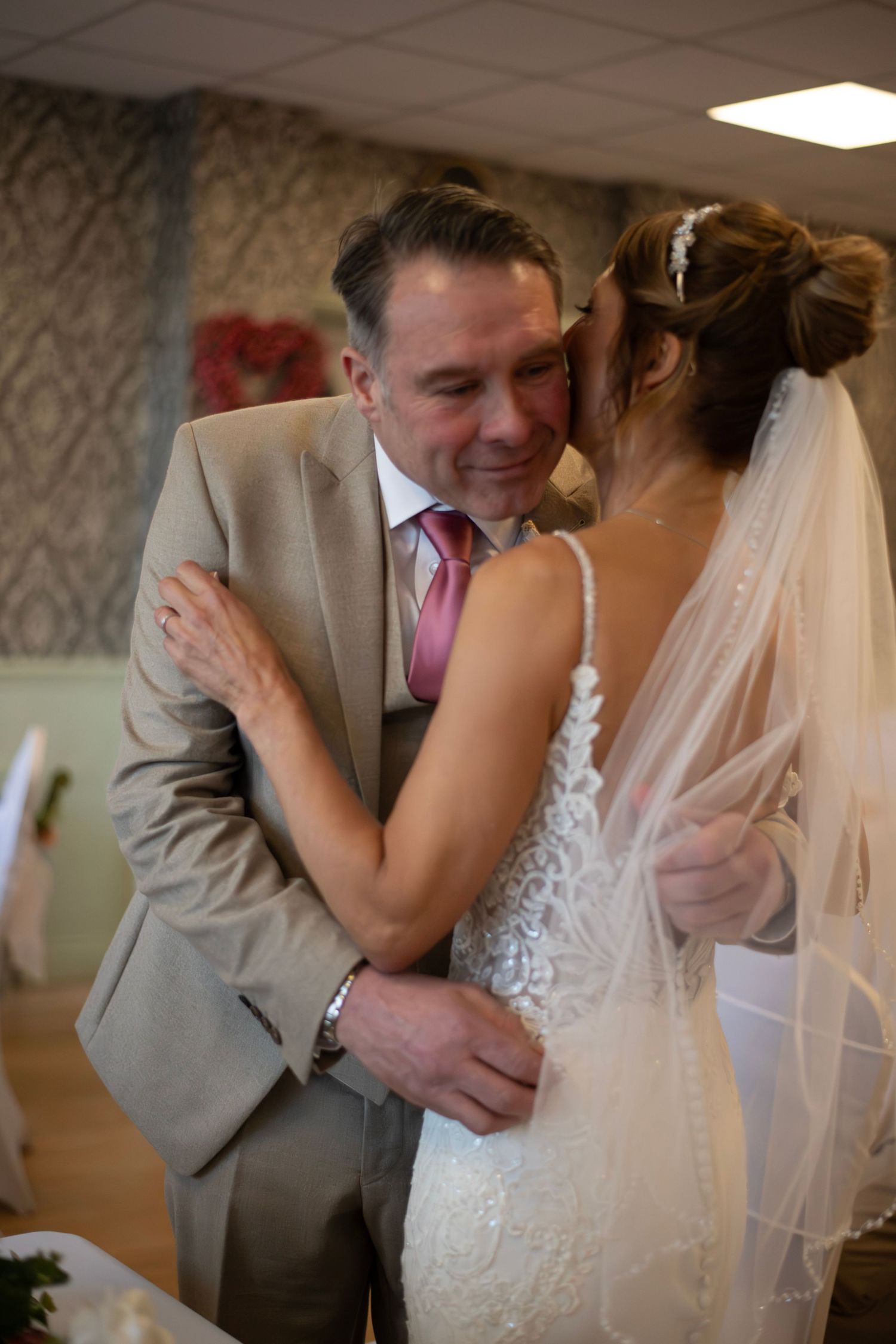 A bride in a lace gown and veil shares an emotional hug with a man in a tan suit and pink tie at a wedding.