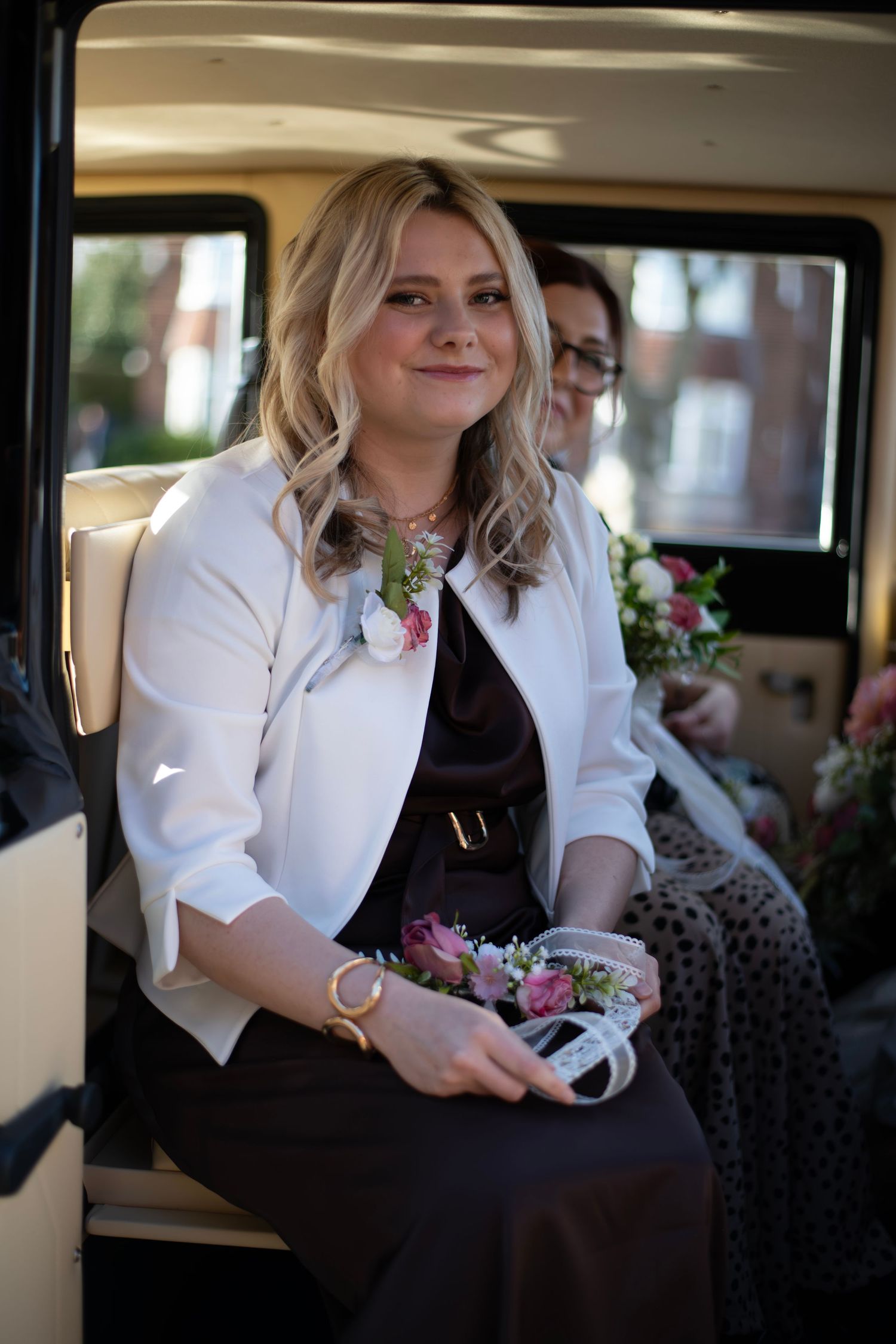 Smiling bridesmaid in white jacket holding floral corsage seated in vintage wedding car.