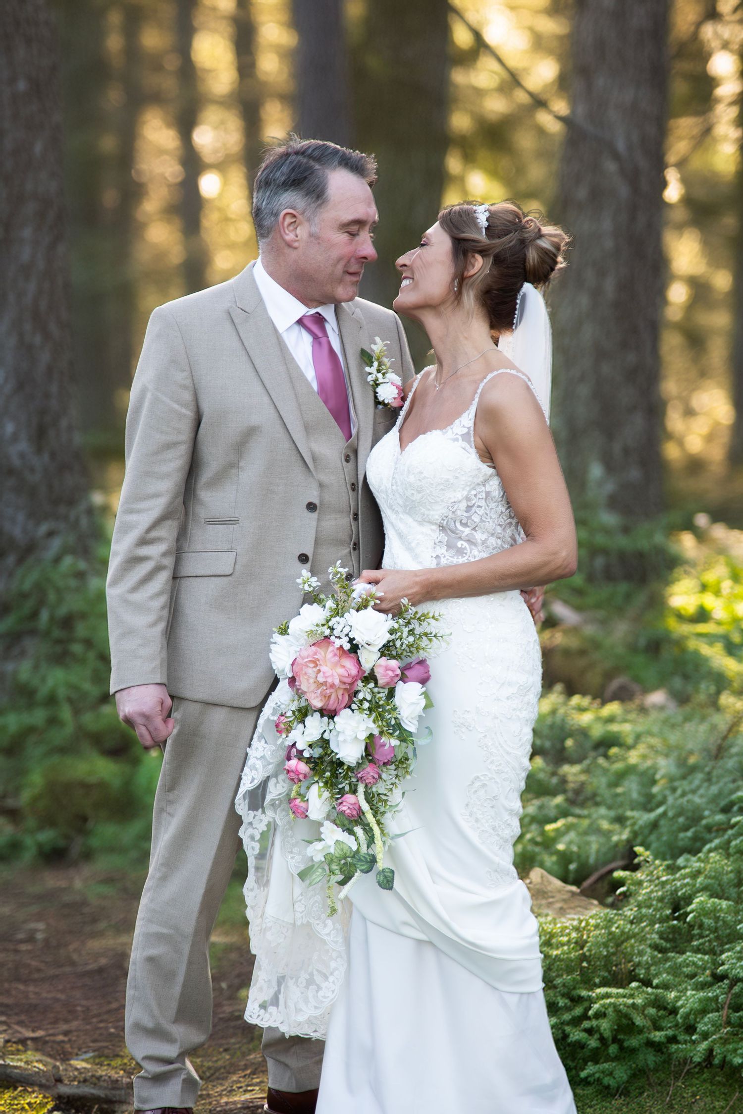 Bride and groom sharing a romantic moment in a forest, bride holding a cascading pink bouquet.