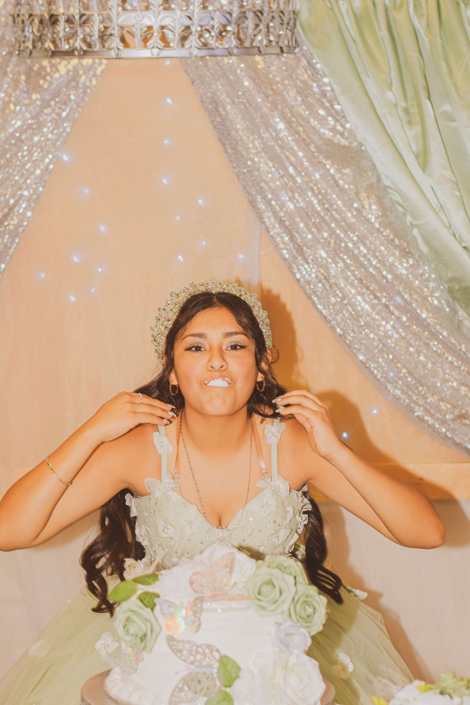 Young Latina girl in green quinceañera gown and tiara poses playfully near sparkly backdrop with floral cake.