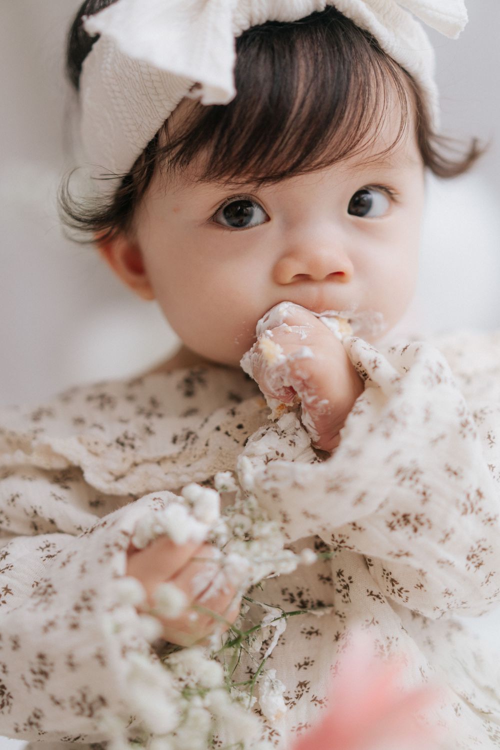 A baby in white lies on floral bedding while looking to the side with a curious expression.一個穿著白色衣服的嬰兒躺在碎花床單上，好奇地看向一旁。