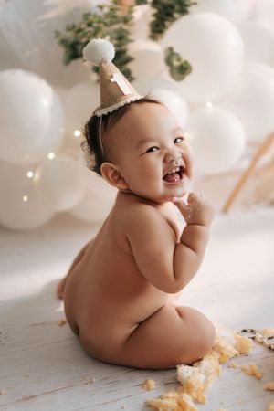 A happy baby in a white bow headband sits on a white floor with cake crumbs during a cake smash photo session.一個戴著白色蝴蝶結髮帶的快樂寶寶坐在鋪滿蛋糕屑的白色地板上，
