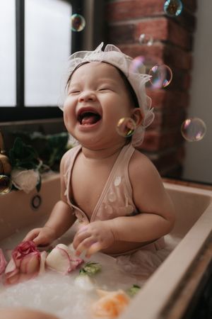 Happy baby laughing in bath with soap bubbles floating around while wearing a beige headband.戴著米色髮帶的快樂寶寶在浴缸裡咯咯笑著，周圍飄著肥皂泡。