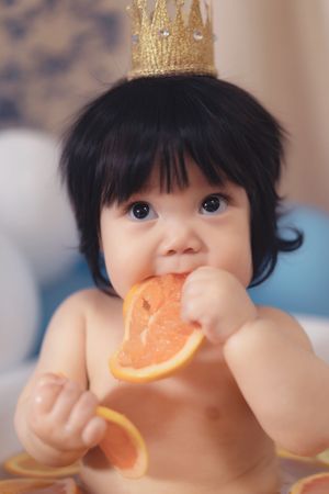A small child wearing a gold crown eats an orange slice while looking at the camera一個戴著金皇冠的小孩一邊看著鏡頭一邊吃著橘子片。
