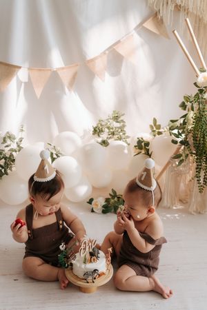 Two babies in birthday hats share a first birthday cake smash surrounded by balloons and boho decorations.兩個戴著生日帽的寶寶在氣球和波西米亞風裝飾的環繞下，一起享用一歲生日