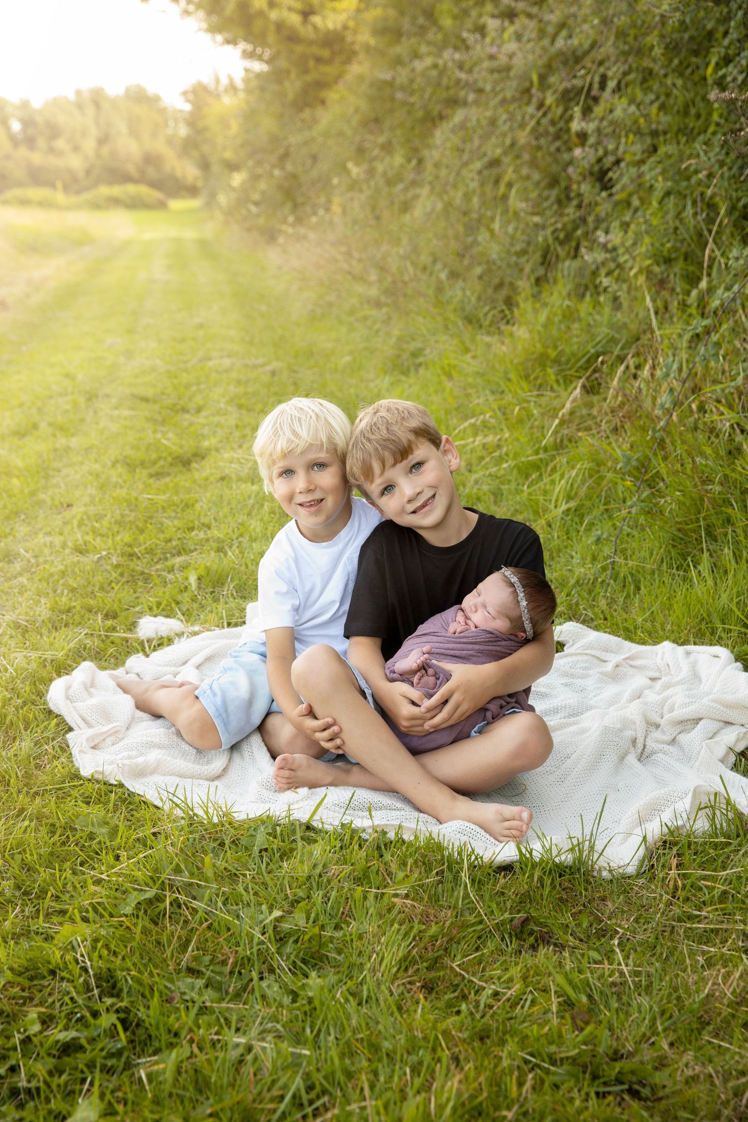 Two blonde boys sit on a white blanket outdoors holding a swaddled newborn baby sibling in golden sunlight. Newborn outdoor family photos.